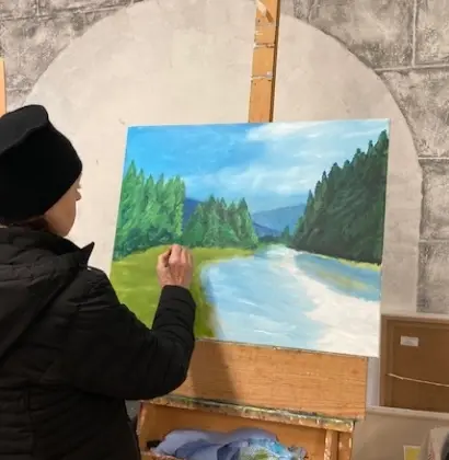 A person in a black jacket and hat paints a landscape on a canvas, featuring a river, trees, and mountains, on an easel indoors. The wall behind has a stone-like arch pattern.