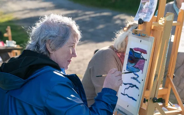 An elderly person with gray hair and a blue jacket paints on paper at an easel outdoors. The artwork features blue and red abstract shapes. Another person in soft focus is painting in the background.
