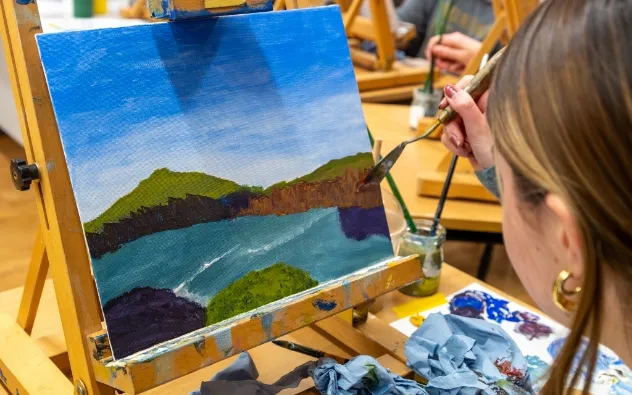 A person painting a landscape on a canvas. The artwork depicts a lake surrounded by hills under a blue sky. The artist holds a brush near the canvas, which is propped on an easel. Various painting materials are visible nearby.