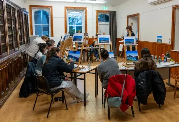 A group of people sit in a classroom, painting landscapes. Canvas paintings of mountains on easels are in front of them. A woman stands near a whiteboard, instructing. The room has wooden floors and walls with bookshelves.