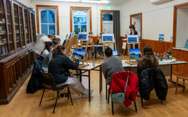 A group of people sit in a classroom, painting landscapes. Canvas paintings of mountains on easels are in front of them. A woman stands near a whiteboard, instructing. The room has wooden floors and walls with bookshelves.