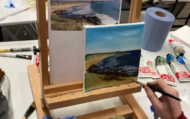 A person is painting a coastal landscape on a canvas. An easel holds both the canvas and a reference photo of a beach. Various paint tubes and brushes are on the table. The painting depicts a beach with grass, ocean, and sky.