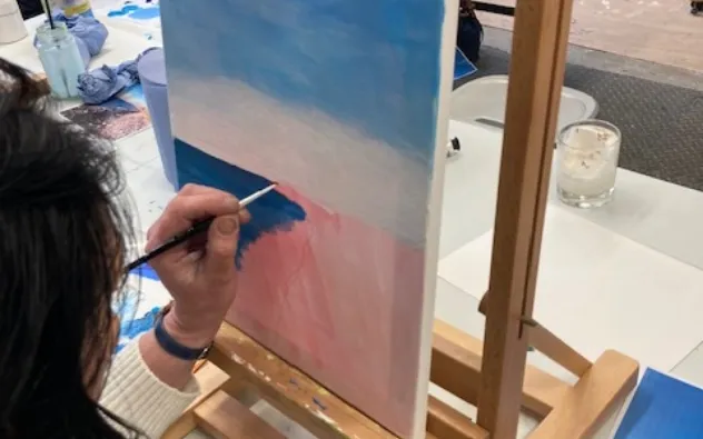 A person painting on a canvas with a brush, creating a landscape of blue sky and pink ground. The canvas is on an easel, and a candle and paint supplies are visible on the table.