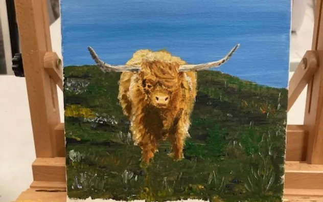A small painting of a Highland cow with long horns and shaggy fur standing on a grassy hill under a blue sky, displayed on a wooden easel.