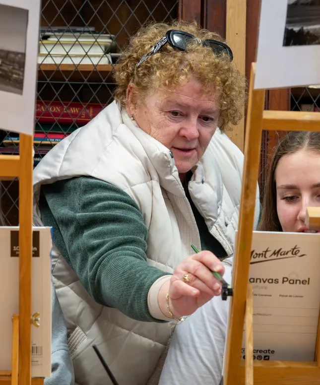 An elderly woman with curly hair, wearing a puffy white vest and green sweater, is painting on a canvas. A younger person beside her looks at the canvas. Shelves with books are visible in the background.