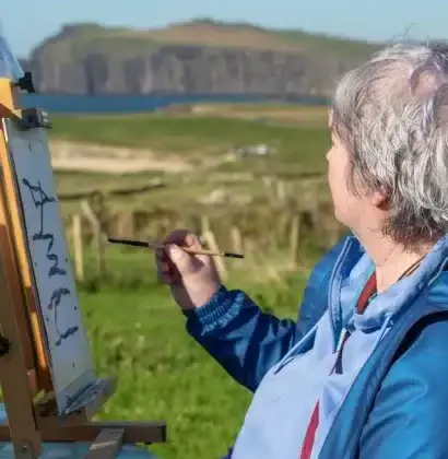 An older person with gray hair is painting on an easel outdoors. They are wearing a blue jacket and are seated on grass. In the background, there is a coastline with cliffs and green fields under a clear blue sky.