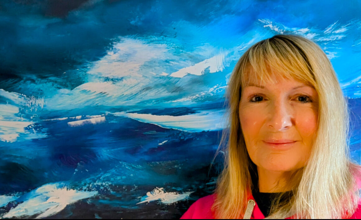 A woman with long blonde hair, wearing a pink top, smiles in front of a vibrant blue and white abstract painting inspired by ocean scenes, capturing the creative spirit of Visiting Artists in Dingle.