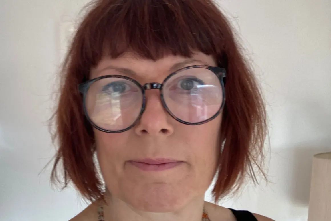 A person with short auburn hair, wearing large round glasses, a sleeveless black top, and a beaded necklace stands indoors with a neutral expression—perhaps one of the Visiting Artists in Dingle. A lamp and cream-colored wall appear in the background.