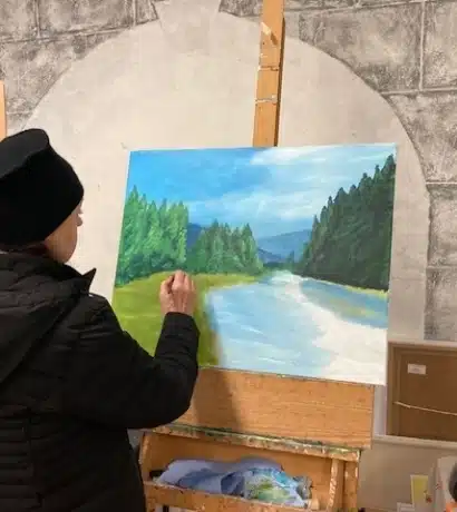 A person wearing a black jacket and hat paints a scenic landscape inspired by Dingle Art, featuring trees, mountains, and a river on a canvas set on an easel indoors.