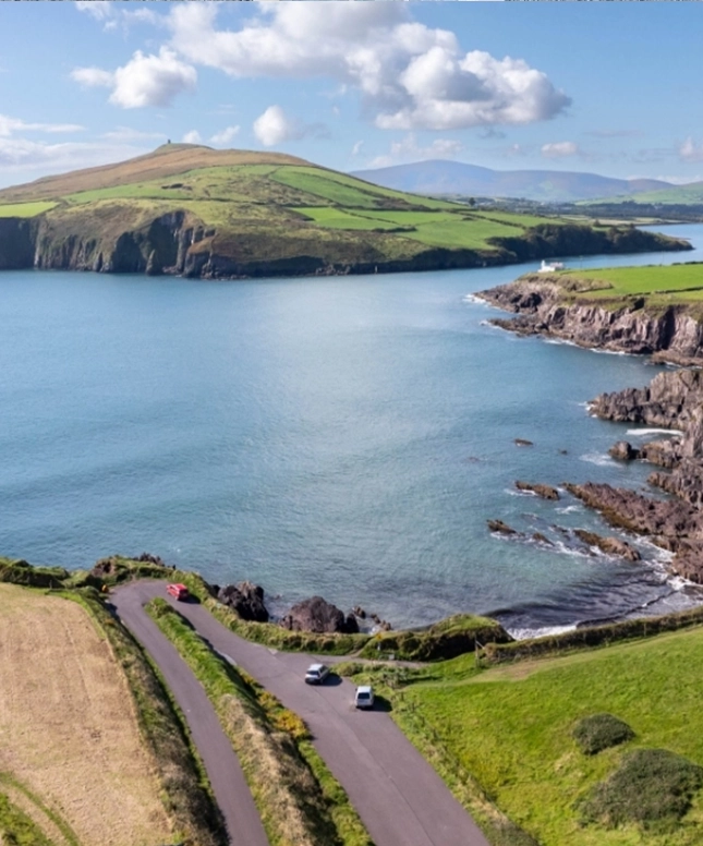 A winding coastal road runs along a rocky shoreline with two parked cars, overlooking calm blue water and green hills under a partly cloudy sky. Visit Dingle for Art and experience inspiring views at every turn.